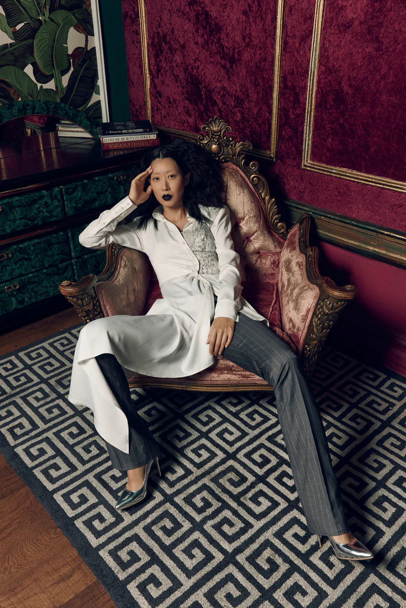A woman with dark lipstick reclines confidently in an ornate velvet armchair, wearing a white asymmetrical shirt paired with gray pinstripe trousers and metallic heels, set against a richly decorated room with red velvet walls and patterned carpet.