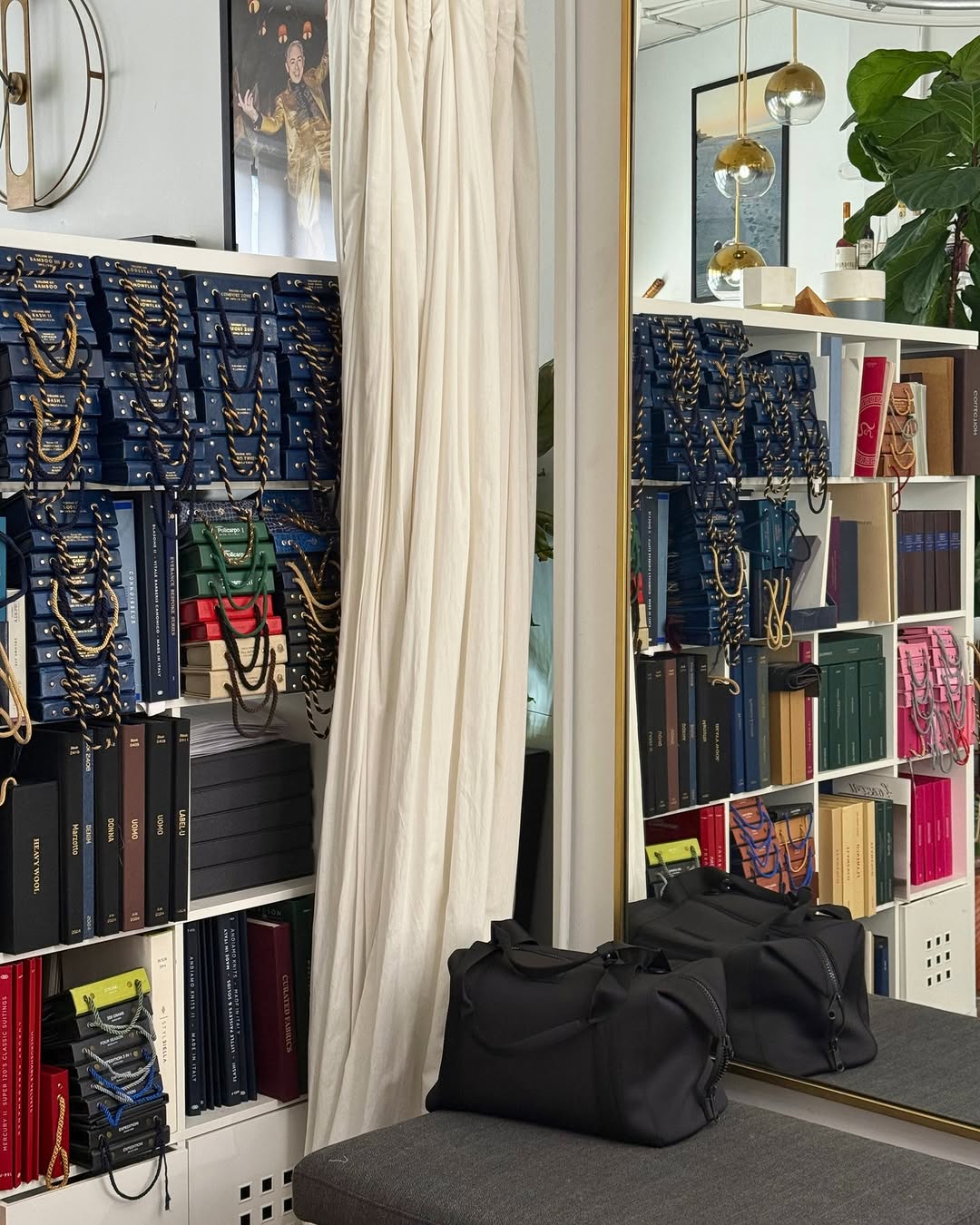 A well-organized shelf unit filled with neatly arranged binders and sample swatches with hanging chains, reflected in a large gold-framed mirror beside a tall white curtain and a black duffel bag on a gray bench.