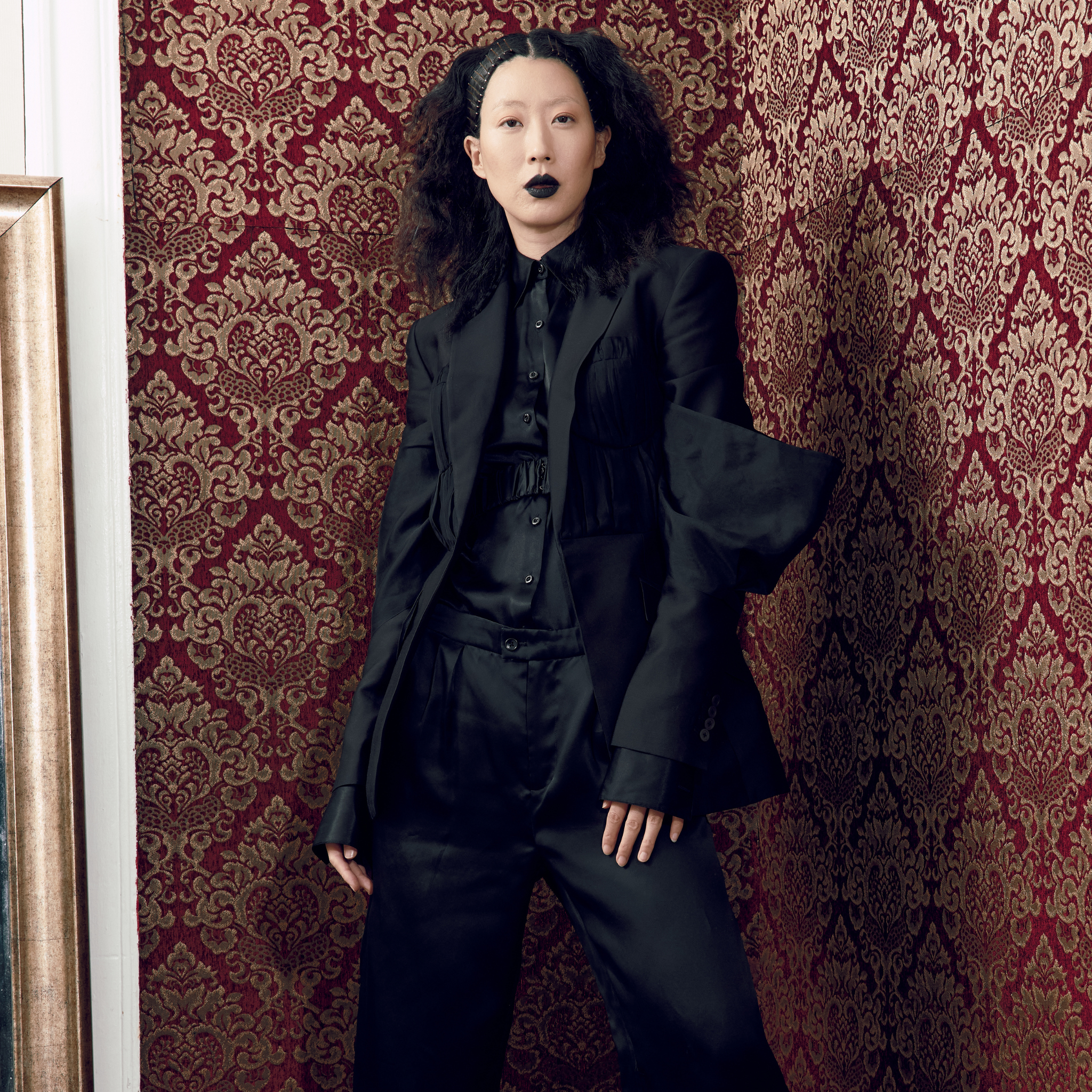Model wearing a black tailored blazer with subtle corset seam detailing and sculpted sleeves, paired with matching trousers and shirt, photographed indoors against a burgundy patterned wall.
