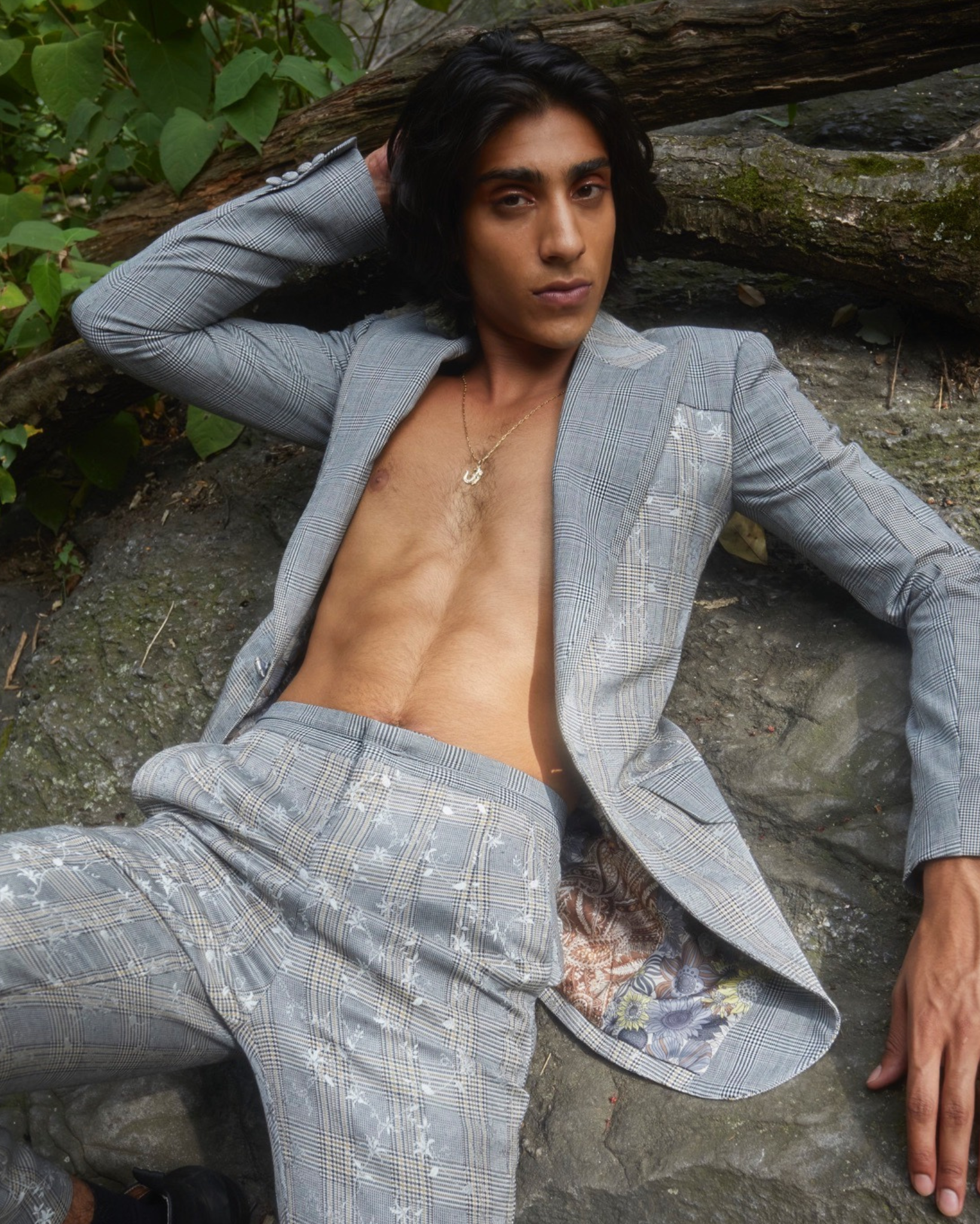 Model wearing a light grey floral plaid tailored blazer with deep V neckline, paired with matching trousers, photographed outdoors reclining on stone and tree roots in a wooded setting.