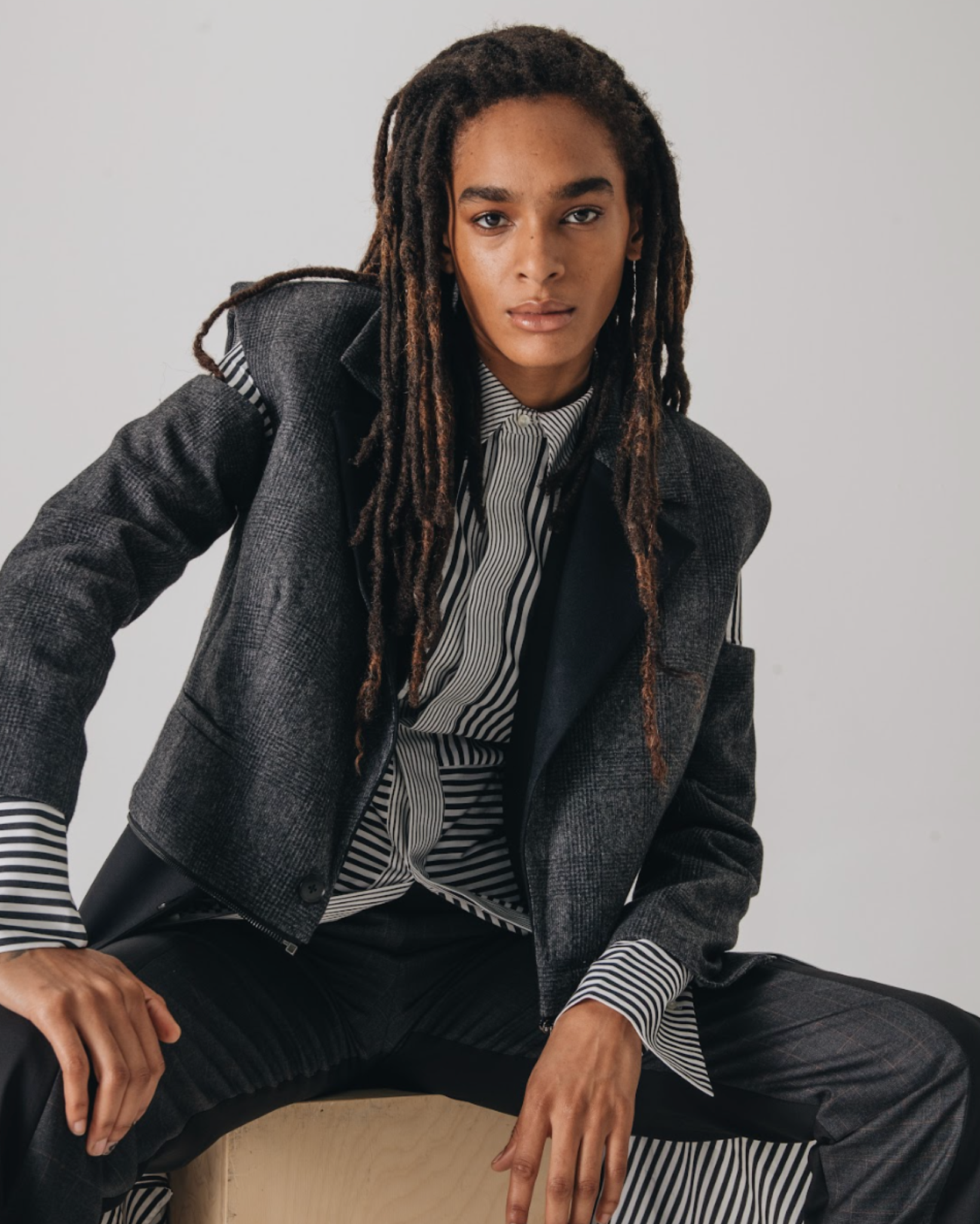 Model wearing a charcoal tailored blazer layered over a black and white striped button up shirt with extended cuffs, paired with black trousers, photographed against a light studio background.