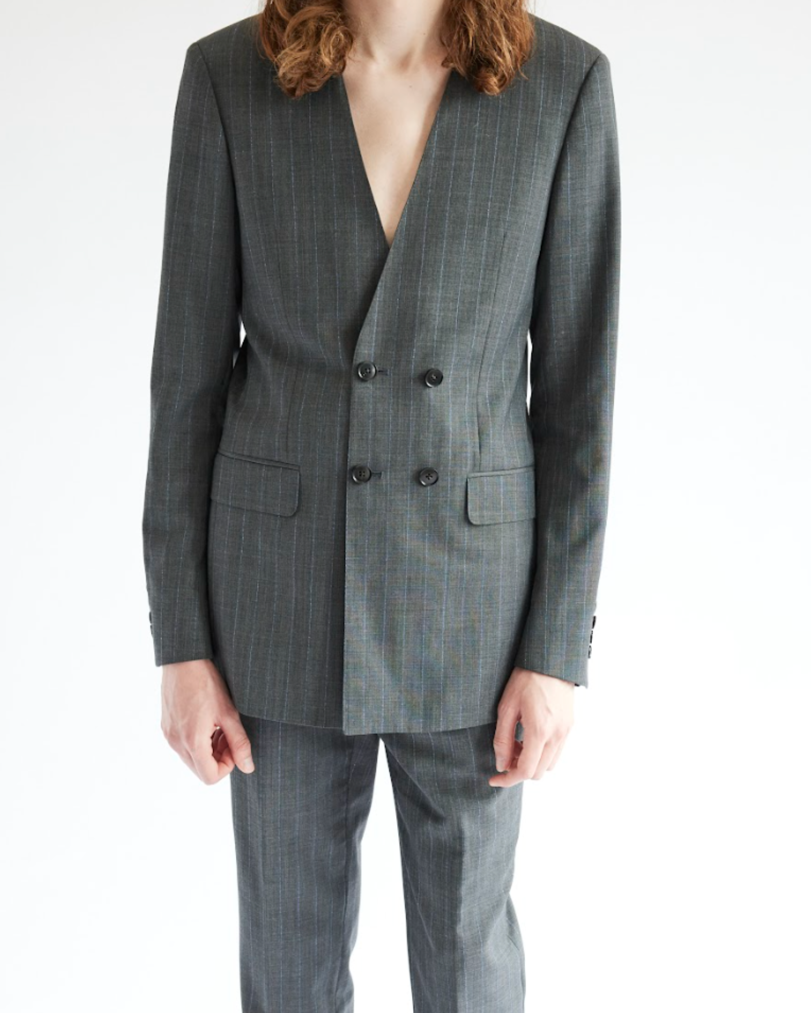 Model wearing a grey double breasted tailored blazer with a deep V neckline and subtle pinstripe texture, paired with matching trousers, photographed against a white studio background.