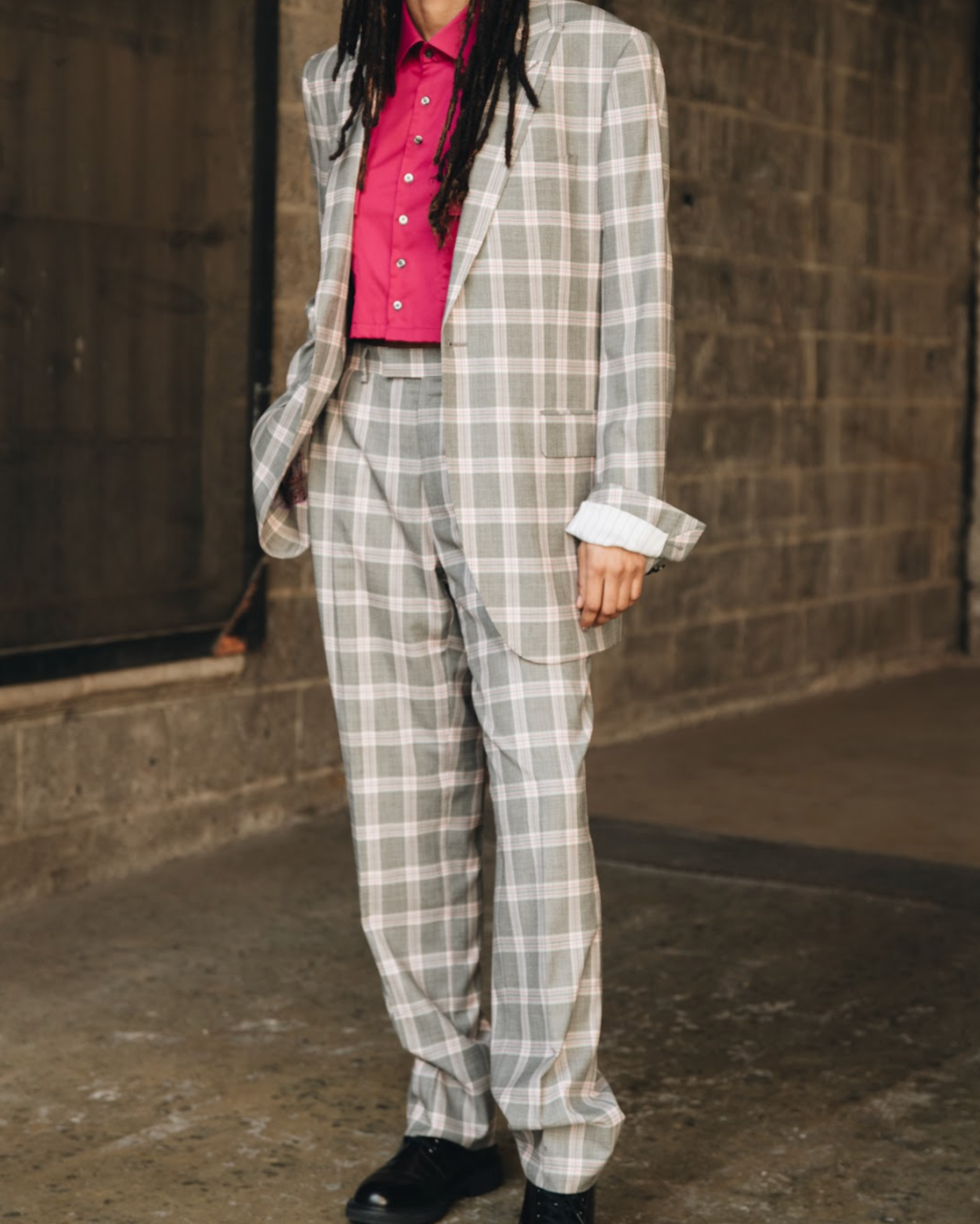 Model wearing a grey plaid tailored blazer with matching trousers and a cropped pink button up shirt, photographed outdoors against a concrete wall background.