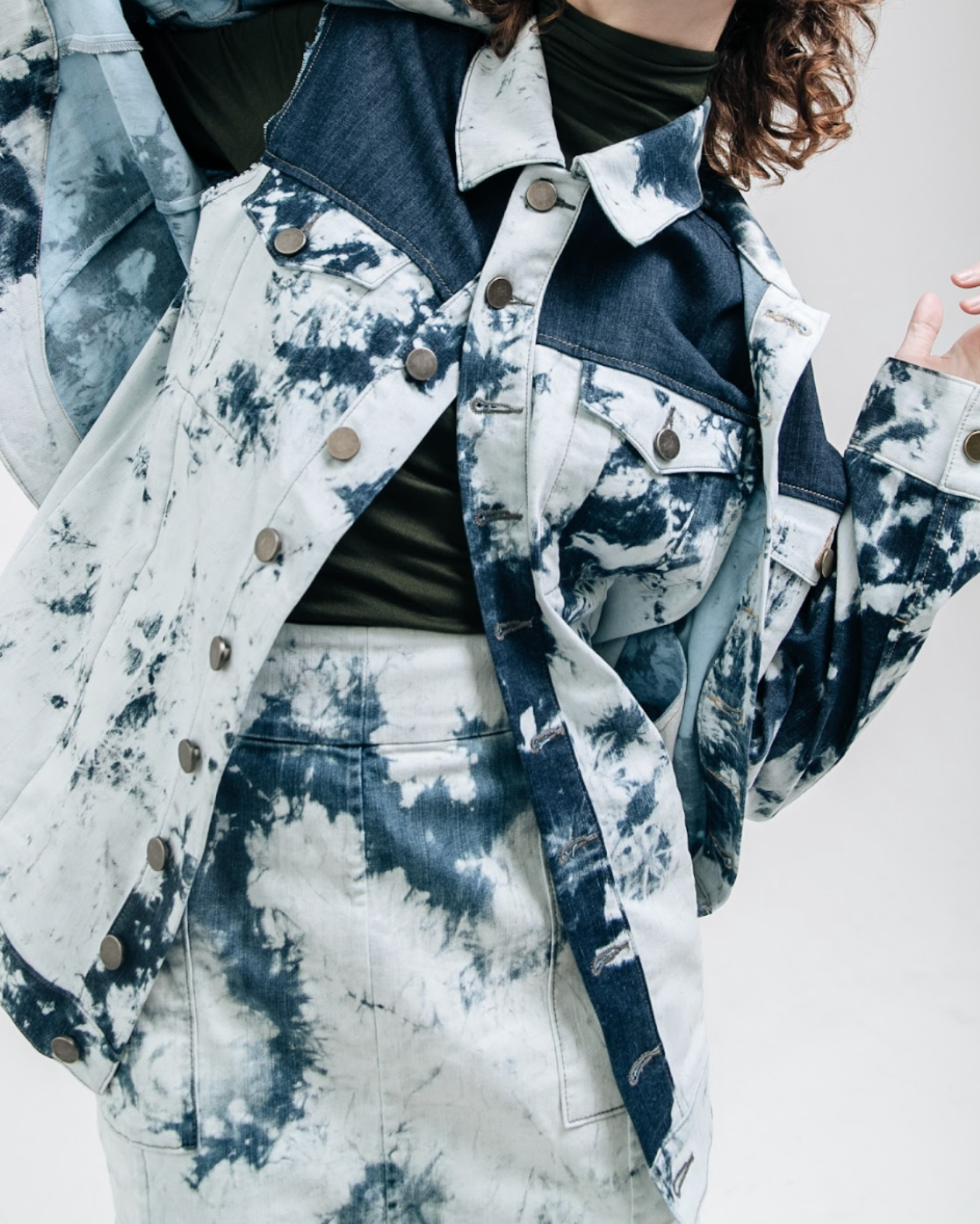 Model wearing an indigo bleached denim jacket with contrast paneling and metal button closure, layered over a dark green top and matching bleached denim skirt, photographed against a white studio background.