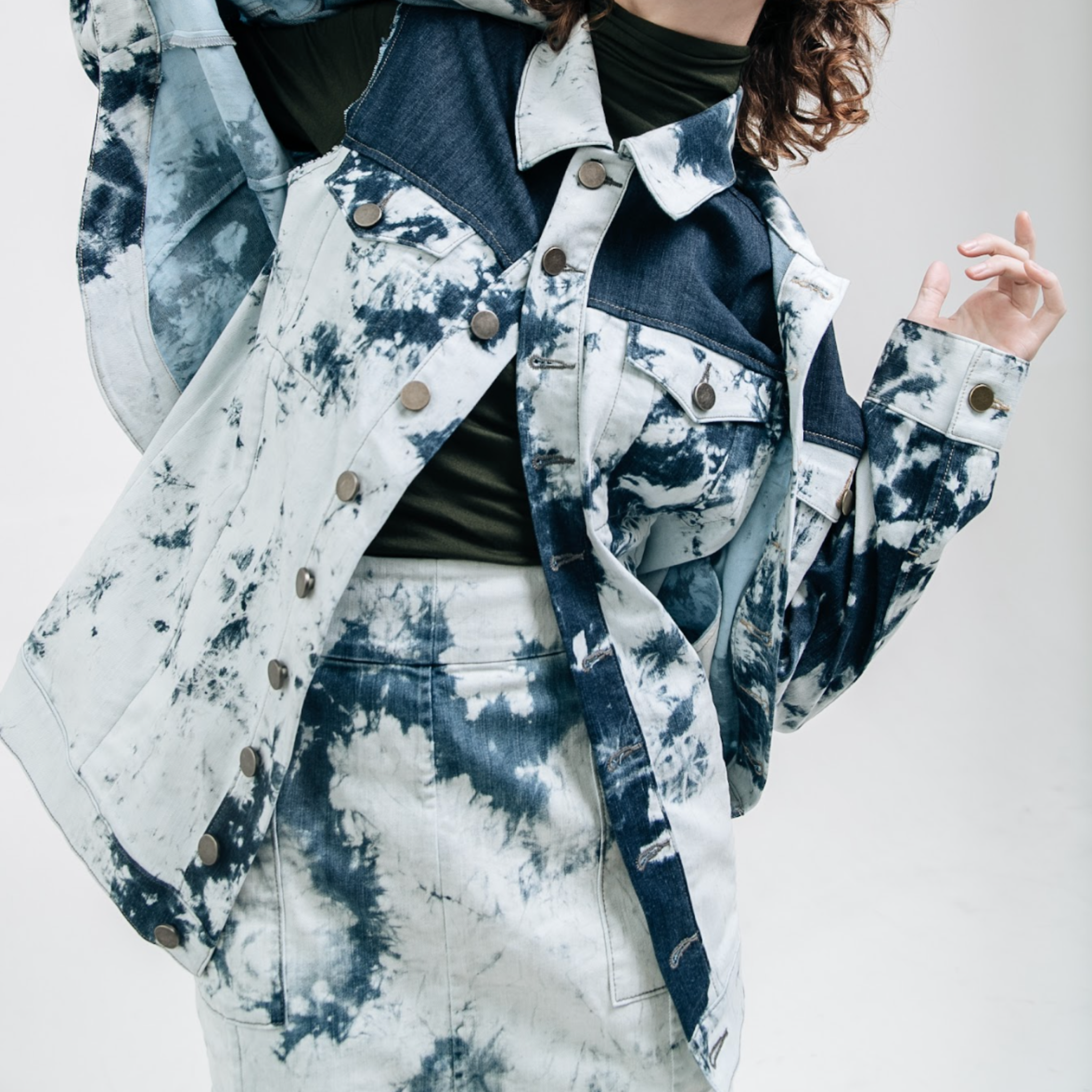 Model wearing an indigo bleached denim jacket with contrast paneling and metal button closure, layered over a dark green top and matching bleached denim skirt, photographed against a white studio background.