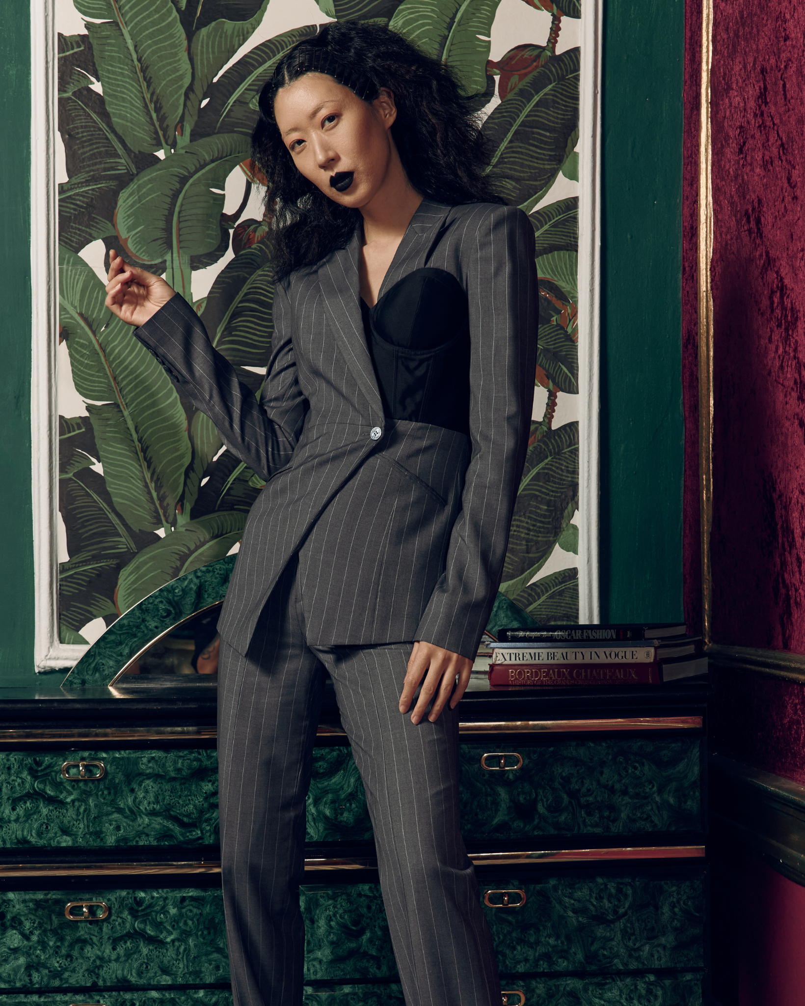 Model wearing a charcoal pinstripe tailored blazer with asymmetric front closure and integrated black corset detailing, paired with matching trousers, photographed indoors in a vintage styled room with patterned wallpaper and rich green and burgundy accents.