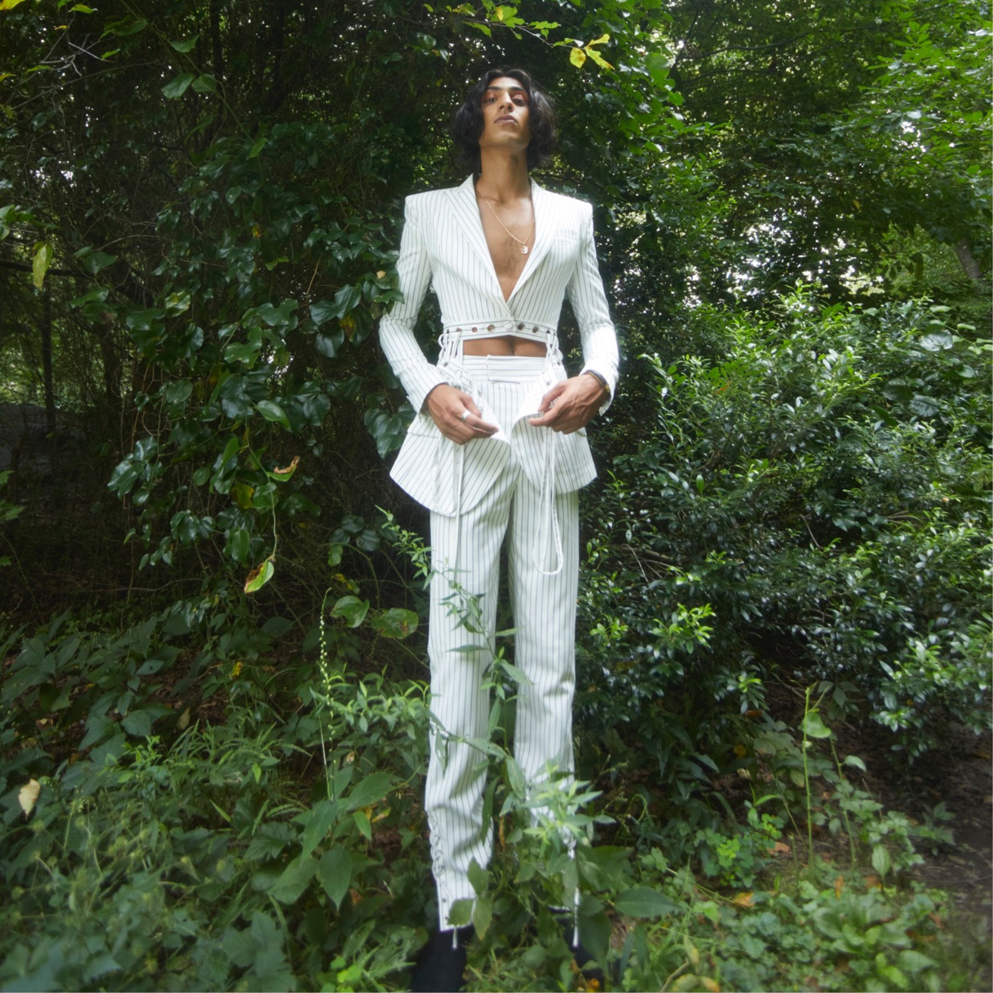 Model wearing a white pinstripe tailored blazer with deep V neckline and waist cutout detailing, paired with matching trousers, photographed outdoors in a wooded setting.