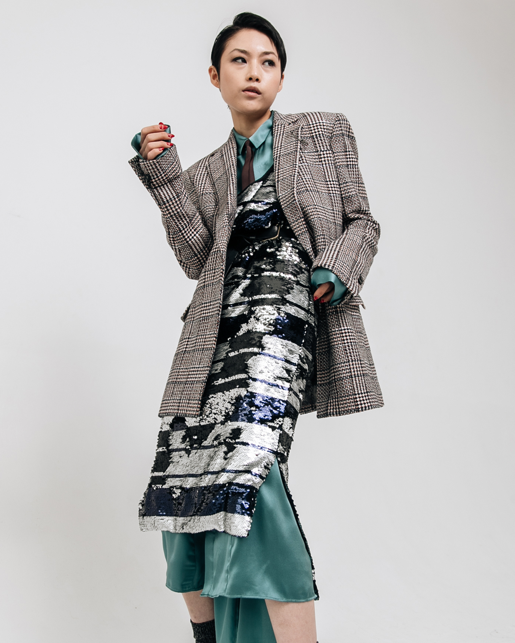 Model wearing a brown and black plaid tailored blazer layered over a silver and navy sequin dress with teal satin underlayer, photographed against a white studio background.