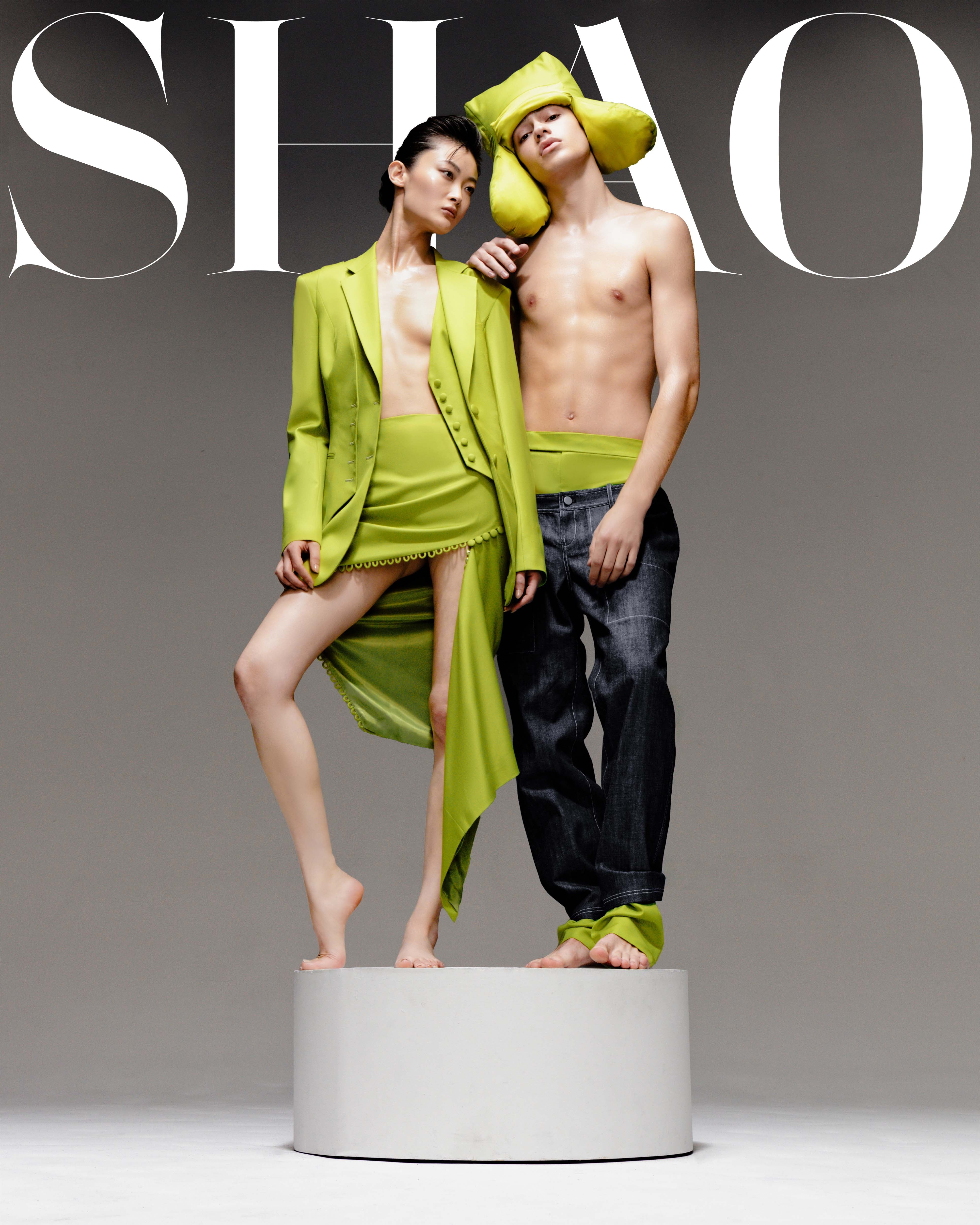 Two models stand barefoot on a round white platform against a neutral backdrop; one wears a lime green asymmetrical skirt and blazer, the other sports dark pants with a matching oversized lime green headpiece.