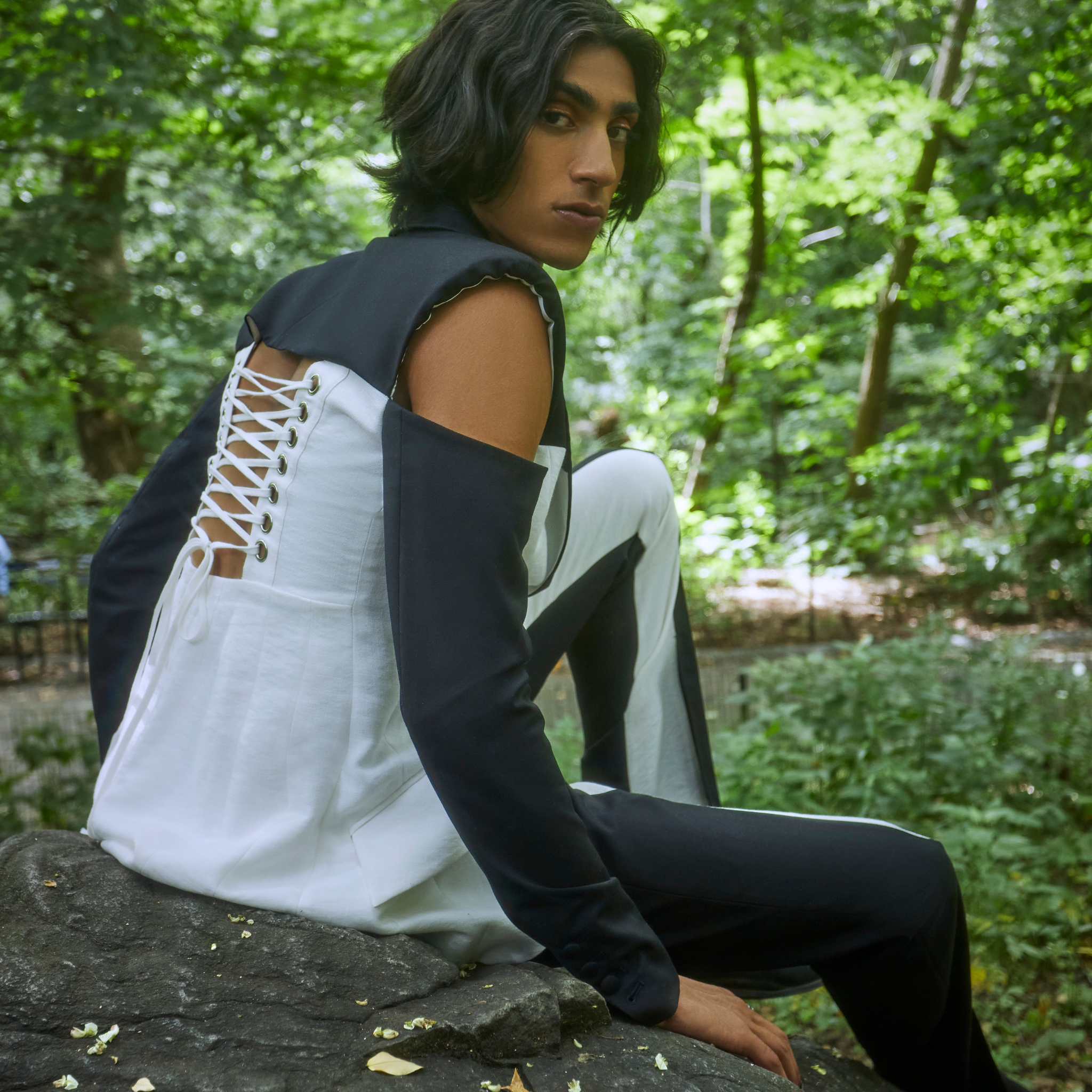 Model wearing a two tone black and ivory tailored blazer with cutout shoulders and lace up back detailing, paired with matching trousers, photographed outdoors in a wooded setting.