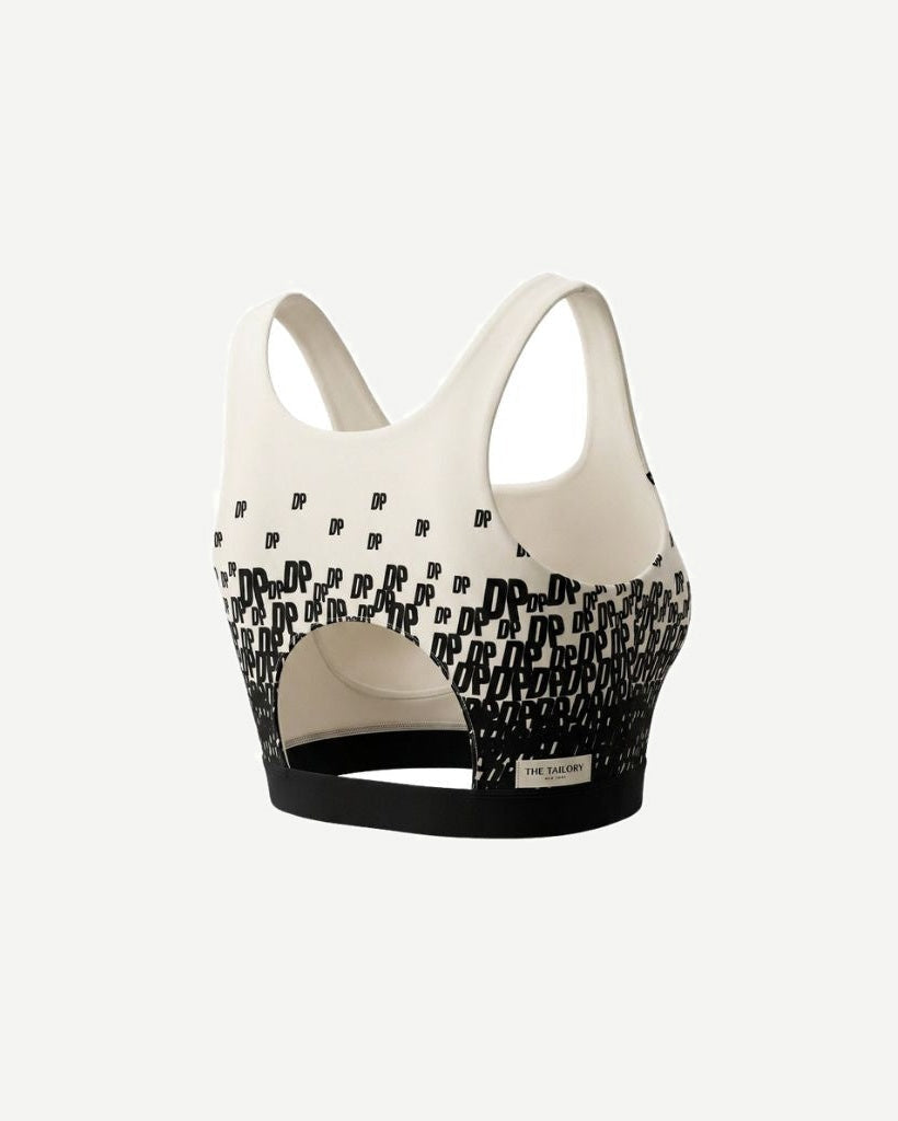 The Tailory New York X Dogpound Cropped Yoga Top in ivory with a black gradient "DP" logo pattern, featuring wide shoulder straps, a cut-out back detail, and a supportive black elastic hem band.