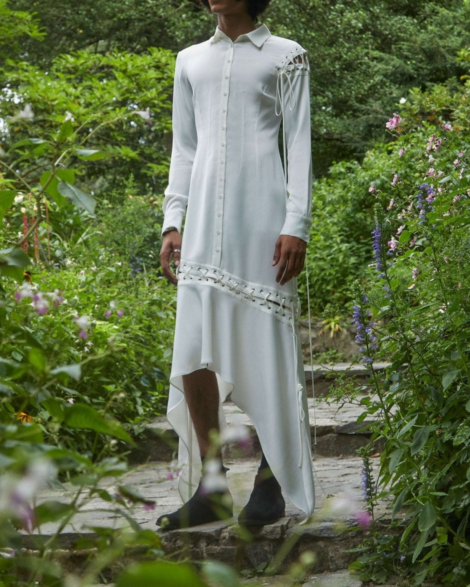 White Cut-Out Shirtdress featuring long sleeves, asymmetric hem, and intricate lace-up cut-out details on shoulder and skirt, styled in a natural garden setting.