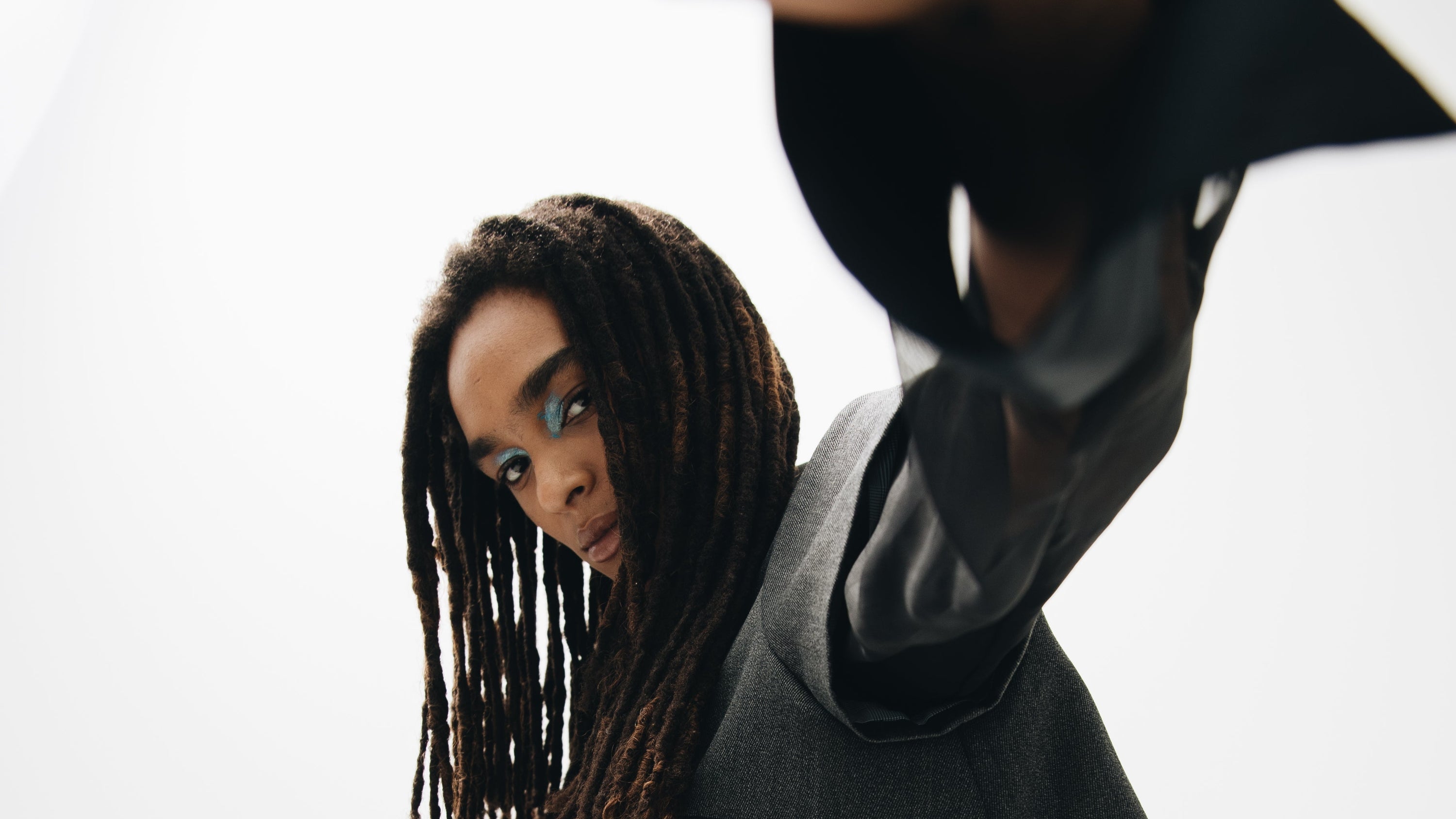 A person with long dreadlocks and blue eyeshadow poses with one arm extended, wearing a sheer black outfit, gray vest, and The Tailory New Yorks Charcoal Silk Flowing Skirt | Liquid Drape against a plain white background.