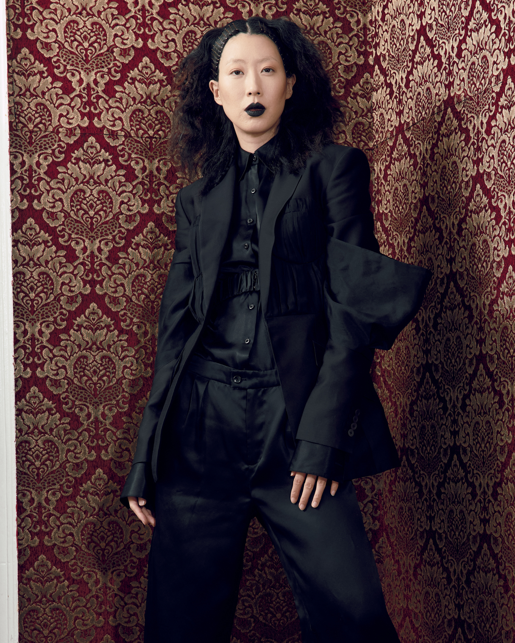 Model wearing a black tailored blazer with subtle corset seam detailing and sculpted sleeves, paired with matching trousers and shirt, photographed indoors against a burgundy patterned wall.
