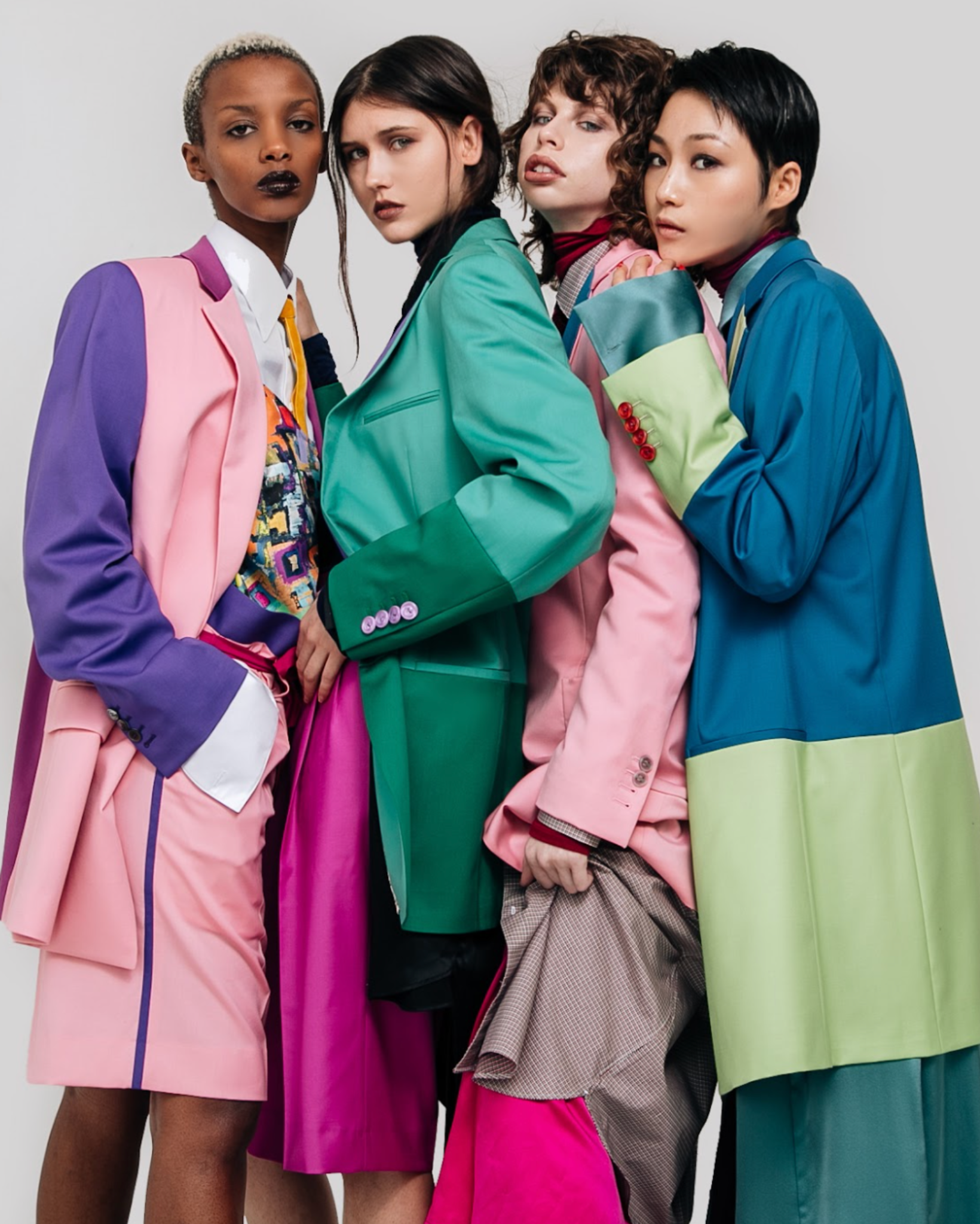 Four models wearing colorblock tailored blazers in pink, purple, green, and blue with contrasting panels, styled with coordinated tailoring pieces, photographed against a light studio background.