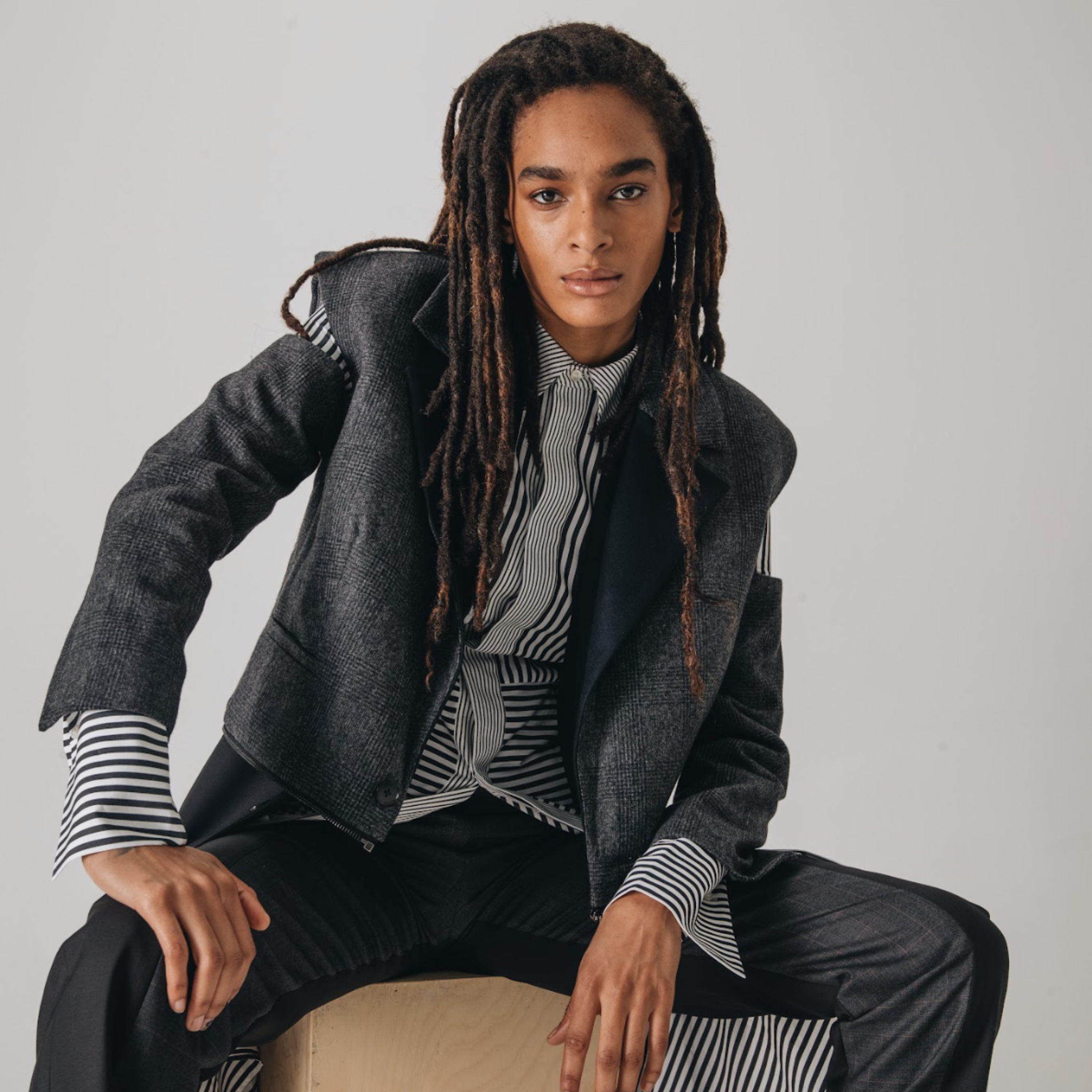 Model wearing a charcoal tailored blazer layered over a black and white striped button up shirt with extended cuffs, paired with black trousers, photographed against a light studio background.