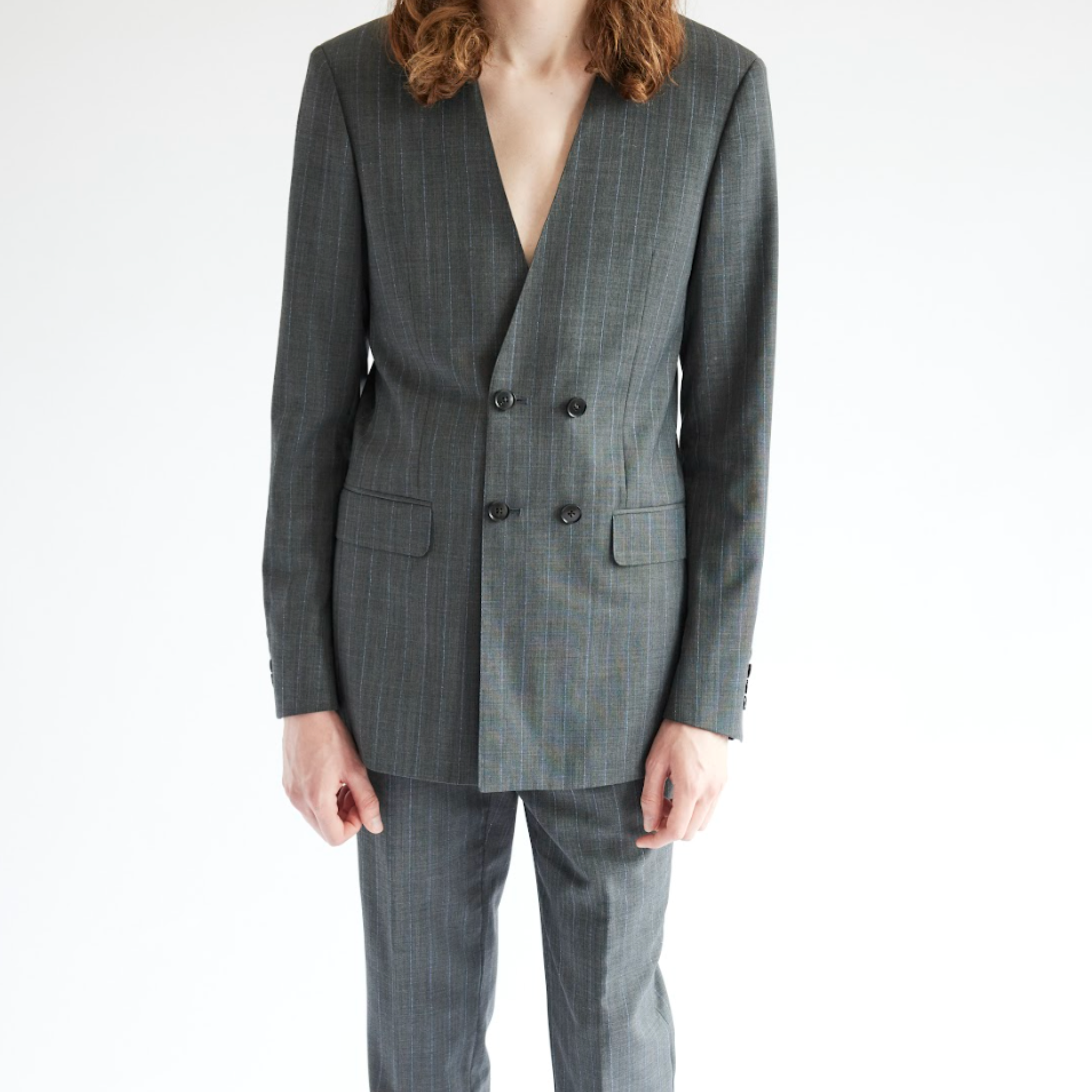 Model wearing a grey double breasted tailored blazer with a deep V neckline and subtle pinstripe texture, paired with matching trousers, photographed against a white studio background.
