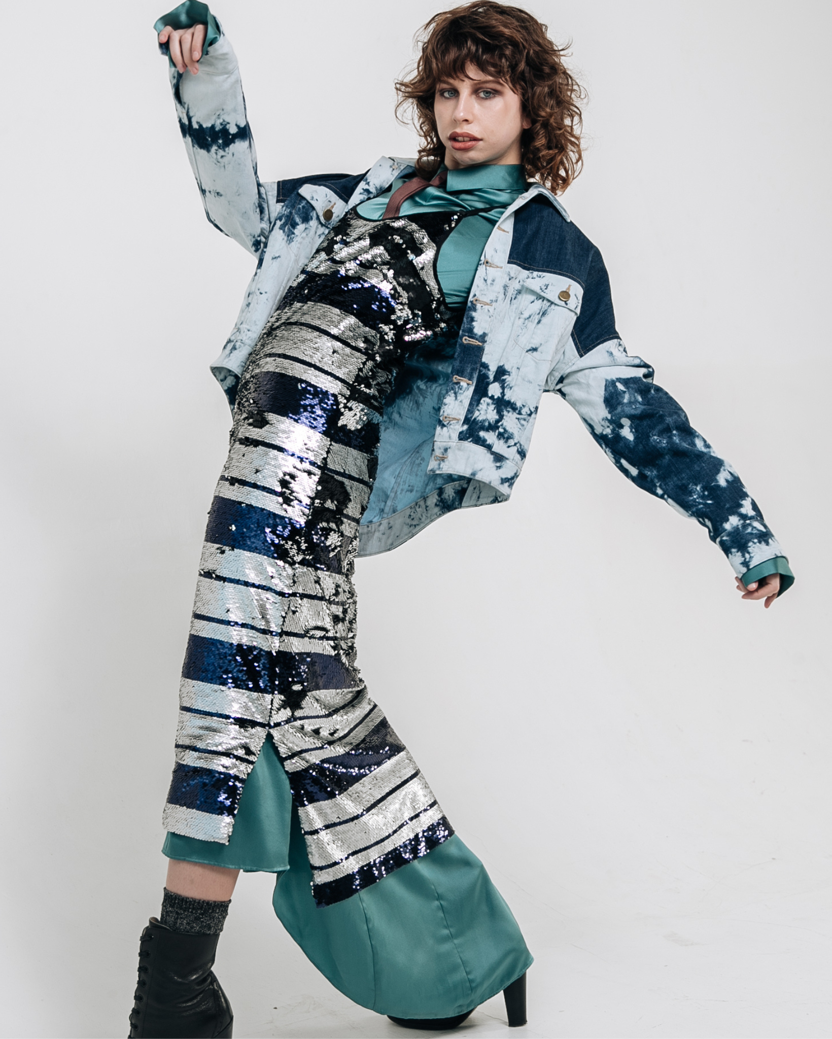 Model wearing an indigo patchwork denim jacket layered over a striped silver and navy sequin midi dress with teal satin underlayer, photographed against a white studio background.
