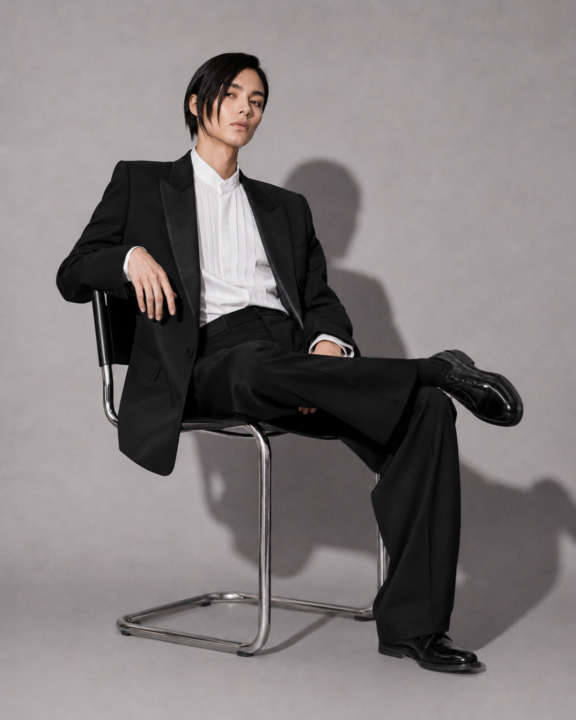 Person wearing a black suit sitting on a chair against a gray background