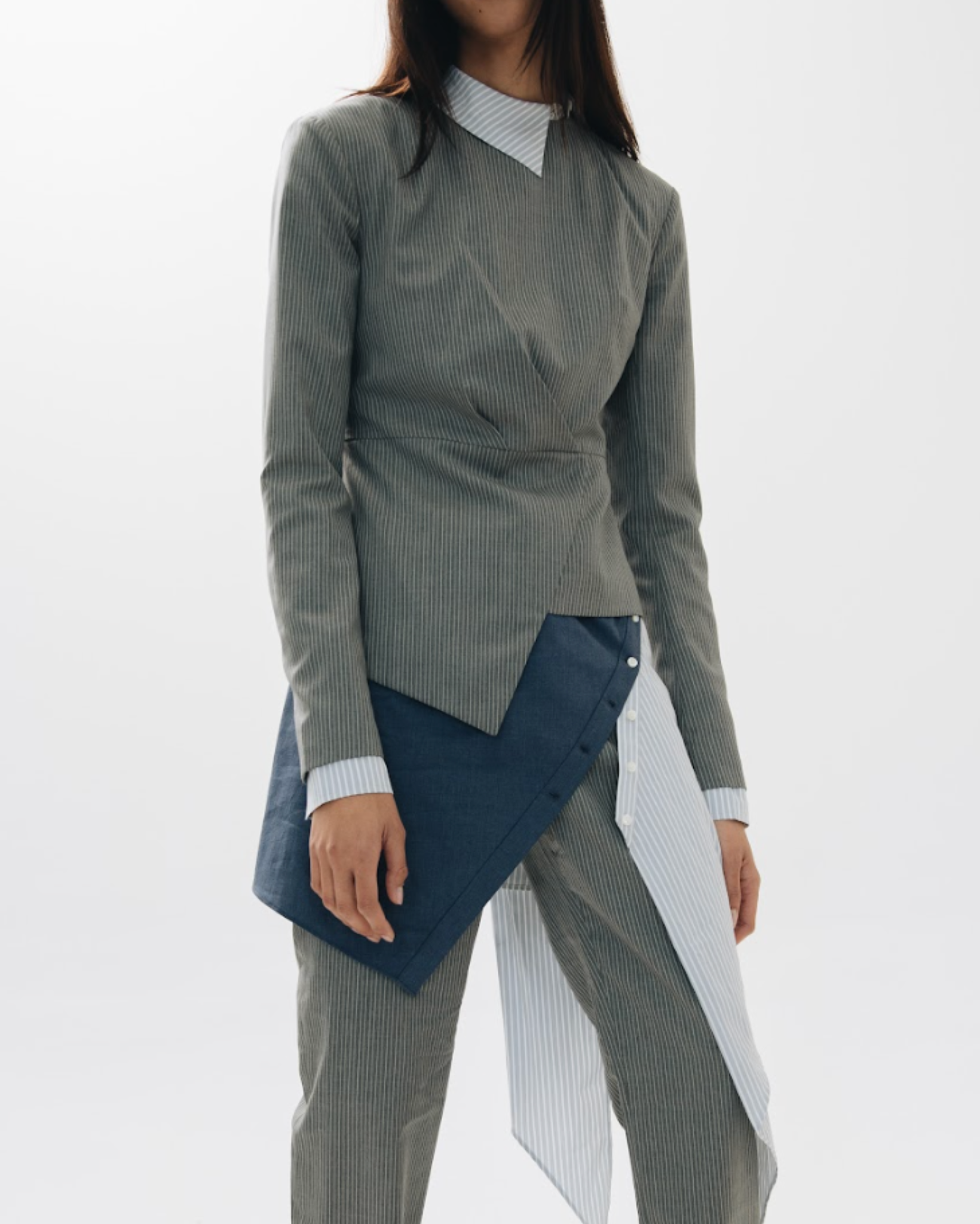 Model wearing the Charcoal Pinstripe Asymmetric Wrap Blouse featuring a subtle vertical pinstripe pattern, a wrap-style front with an uneven hem, and layered shirt detail at the collar and cuffs.