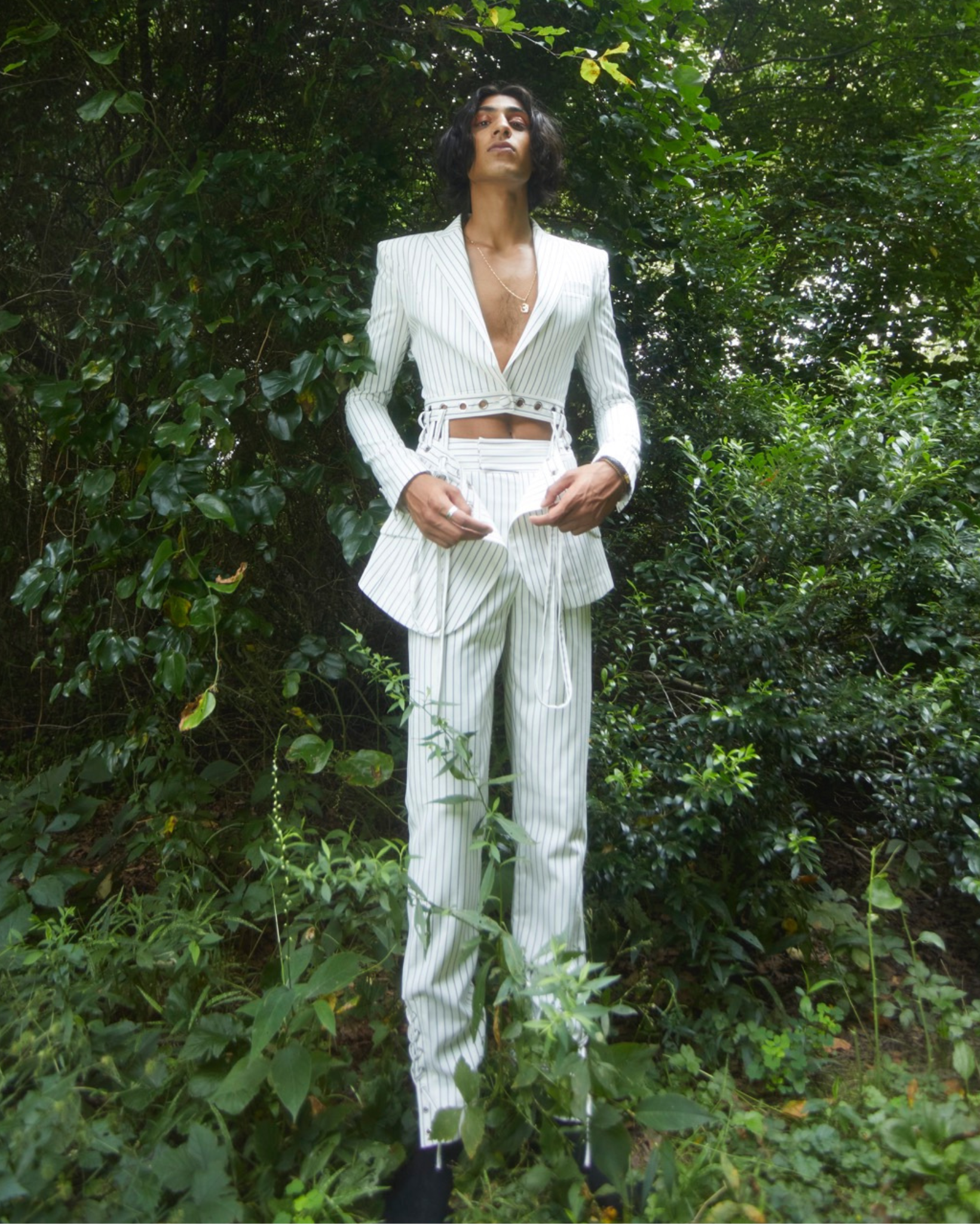 Model wearing a white pinstripe tailored blazer with deep V neckline and waist cutout detailing, paired with matching trousers, photographed outdoors in a wooded setting.