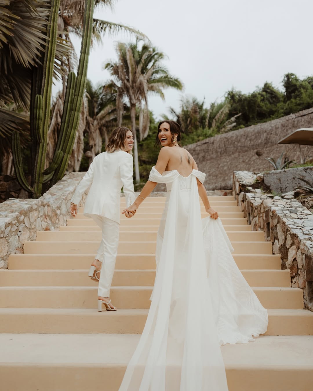 Two women in white outfits walking hand-in-hand on a stone staircase with tropical plants in the background.