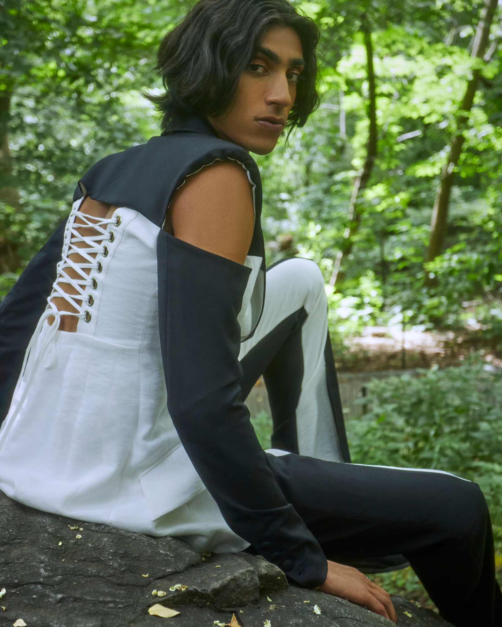 Model wearing a two tone black and ivory tailored blazer with cutout shoulders and lace up back detailing, paired with matching trousers, photographed outdoors in a wooded setting.