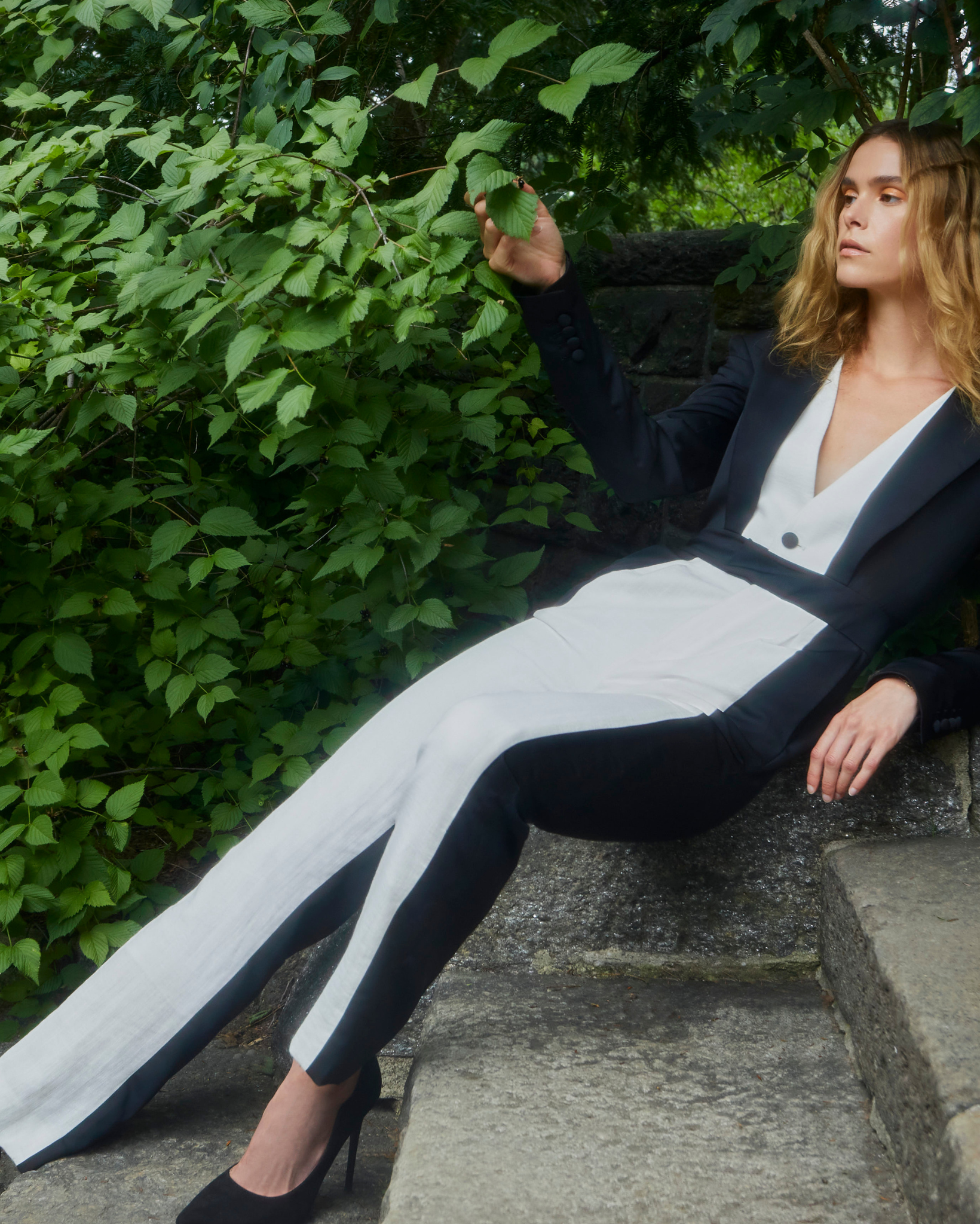 Model wearing two tone black and ivory tailored trousers with contrasting side panels, styled with a matching blazer, photographed outdoors on stone steps surrounded by greenery.