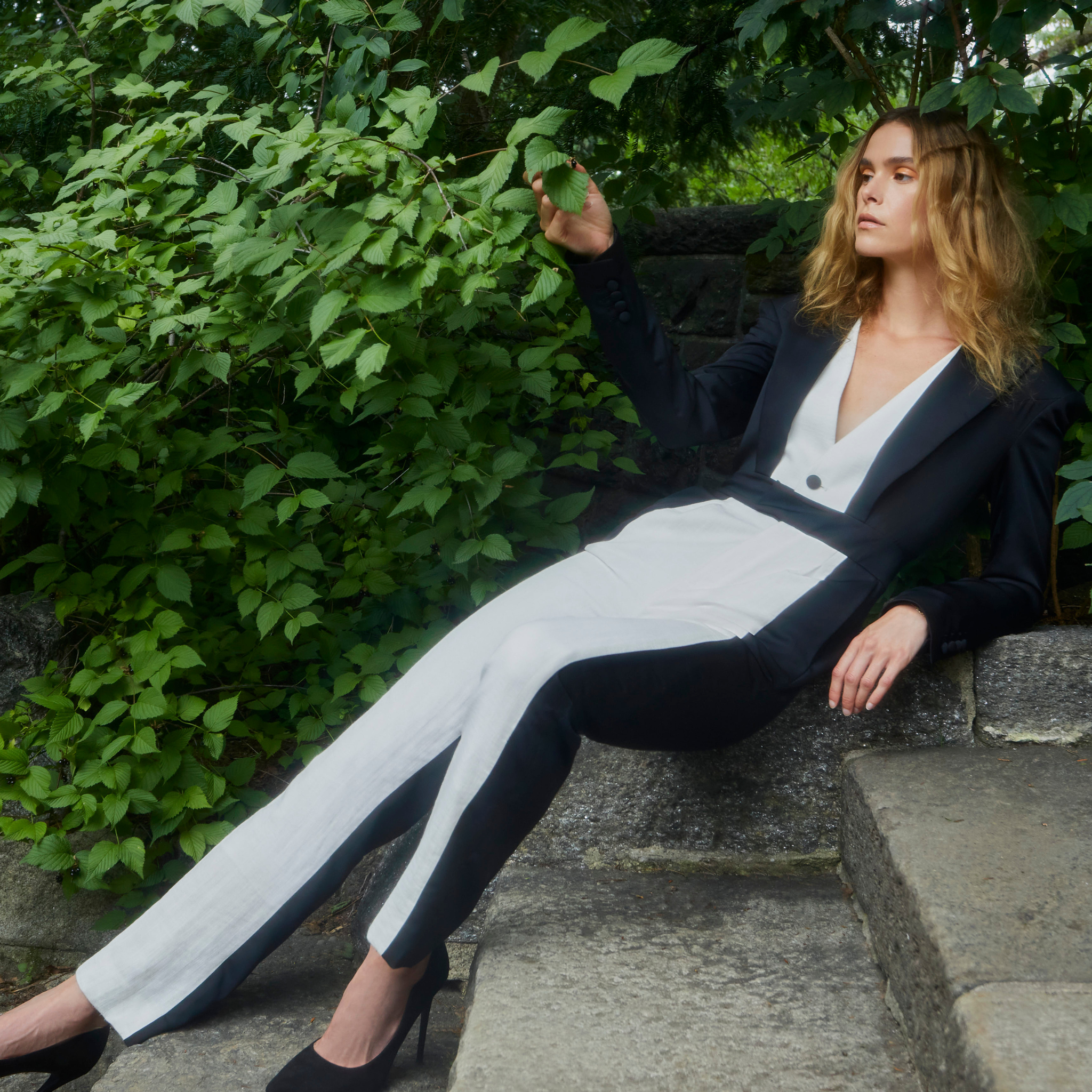 Model wearing two tone black and ivory tailored trousers with contrasting side panels, styled with a matching blazer, photographed outdoors on stone steps surrounded by greenery.