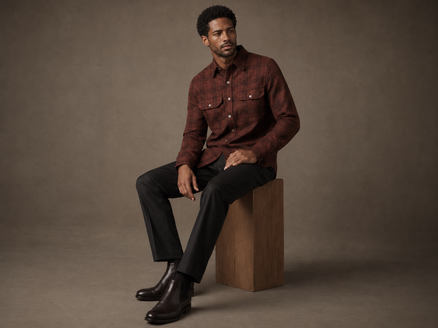 Man sitting on a wooden block wearing a brown plaid shirt and black pants against a brown background